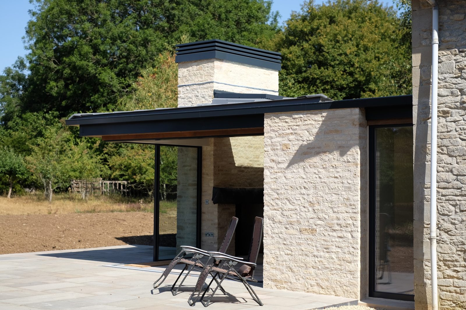 The completed cottage extension in Cirencester: a flat-roofed glazed addition with a stone chimney opening onto a new terrace