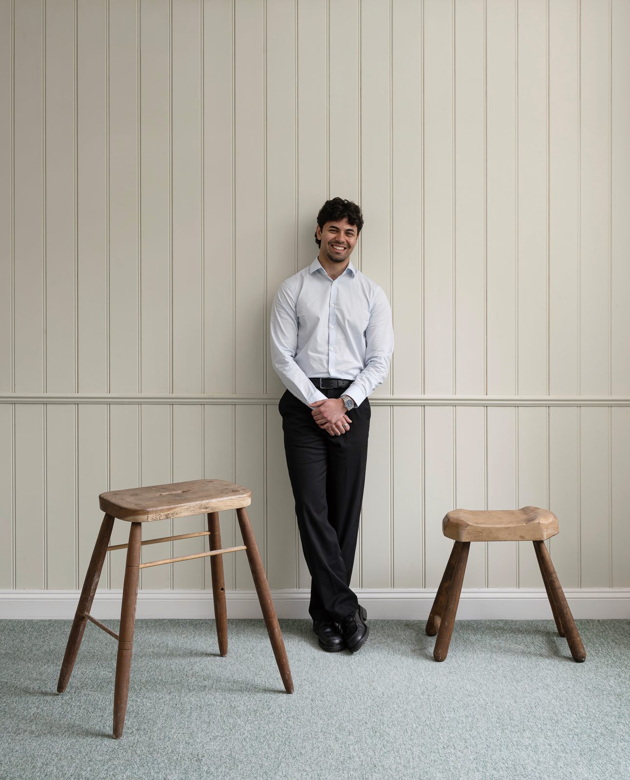 Arnav standing against a panelled wall between two timber stools
